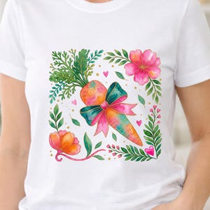 Puede incluir: Camiseta blanca con un diseño de acuarela de una zanahoria con un lazo verde azulado y rosa, rodeada de flores rosas, hojas verdes y pequeños corazones rosas. El diseño tiene una estética caprichosa y primaveral.
