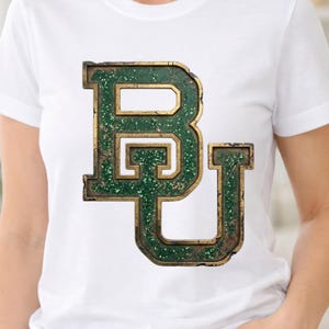 Puede incluir: Camiseta blanca con el logo "BU" verde y dorado desgastado. El diseño universitario tiene un aspecto texturizado y brillante. La camiseta es de estilo cuello redondo clásico.