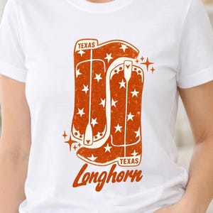 Puede incluir: Camiseta blanca con un diseño gráfico naranja de botas de vaquero y estrellas. El texto "TEXAS" está en la parte superior y "Longhorn" en la inferior, en una fuente estilizada. El diseño tiene un aspecto vintage y desgastado.