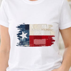 Puede incluir: Camiseta blanca con un diseño de la bandera de Texas. La bandera está compuesta por varios parches de tela, incluyendo mezclilla, encaje y cuadros, creando una apariencia texturizada única. El diseño de la bandera de Texas incluye una estrella blanca sobre un fondo azul, una franja blanca y una franja roja.
