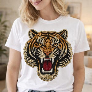 Puede incluir: Camiseta blanca con un diseño detallado de la cara de un tigre. La cara del tigre está representada con cuentas negras, doradas y naranjas, con la boca roja abierta. La camiseta tiene mangas cortas y cuello redondo.