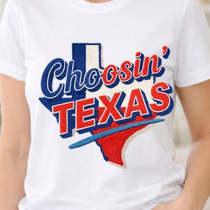 Puede incluir: Camiseta blanca con un diseño bordado que presenta la palabra "Choosin'" en escritura azul sobre la palabra "TEXAS" en letras rojas. El diseño tiene la forma del estado de Texas, con detalles en azul y rojo.