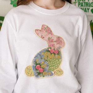 Puede incluir: Camiseta blanca de manga larga con un diseño de conejito de patchwork. El conejito tiene una cabeza floral rosa, cuerpo azul y amarillo y vientre de lunares verdes. Pequeños acentos florales rosas están colocados en el conejito. Estilo cómodo y casual.