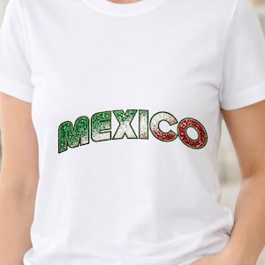 以下が含まれることがあります： 胸元にグリッター調の質感で「MEXICO」という文字がプリントされた白い半袖Tシャツ。文字の色は左側の緑から中央の白、右側の赤へと変化しており、国旗を模したデザインになっています。