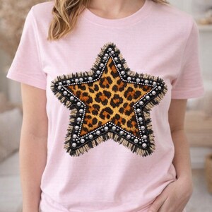 Puede incluir: Camiseta rosa claro con un diseño de estrella grande. La estrella tiene un centro con estampado de leopardo, un borde de flecos negros y está adornada con pequeños tachuelas redondas plateadas. La camiseta es de manga corta.