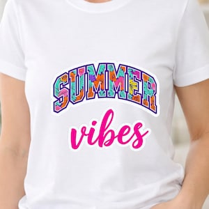 Pode incluir: Uma t-shirt branca de manga curta com o texto SUMMER numa fonte de bloco arrojada com contorno escuro, preenchida com um padrão floral colorido de flores cor de laranja, rosa e turquesa, seguido da palavra vibes em letra cursiva rosa.
