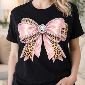 Puede incluir: Camiseta negra con un diseño de lazo grande. El lazo tiene un centro de satén rosa, detalles de estampado de leopardo y está adornado con pedrería. Una gran pedrería está en el centro del lazo.