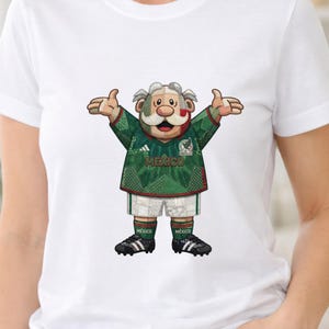 Könnte beinhalten: Ein weißes T-Shirt mit einer Grafik einer Zeichentrickfigur mit Schnurrbart, die ein grünes Mexiko-Fußballtrikot, weiße Shorts und schwarze Fußballschuhe trägt. Die Figur hat die Arme ausgebreitet und ist in einem Patchwork-Muster gestaltet.