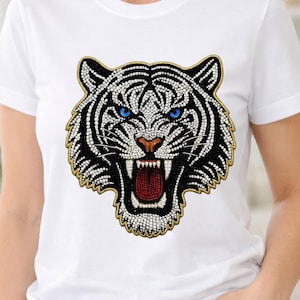 Puede incluir: Camiseta blanca con un diseño de cabeza de tigre adornado con cuentas. El tigre tiene rayas negras y blancas, ojos azules y la boca abierta con la lengua roja. El diseño está adornado con pequeñas cuentas brillantes y contorneado en dorado.