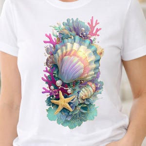 Puede incluir: Una camiseta blanca con un estampado gráfico central de una gran concha iridiscente rodeada de coral rosa, una estrella de mar dorada, follaje acuático azul y pequeñas perlas blancas con una vibrante paleta de colores pastel.