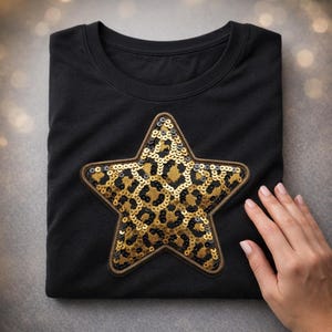 May include: Black t-shirt with a large star-shaped sequin patch. The star features a gold and black leopard print design, outlined with a brown border. The shirt is folded neatly, and a hand is visible on the right side.