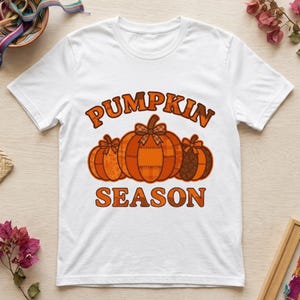 Puede incluir: Una camiseta blanca de manga corta con un gráfico de tres calabazas naranjas con lazos. El texto PUMPKIN SEASON está impreso en letras naranjas y marrones sobre y debajo de las calabazas, incluyendo patrones de cuadros y flores.