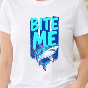 May include: A white short-sleeved t-shirt featuring a graphic print of a shark in shades of blue, pink, and white. The text "BITE ME" is printed in large, light blue letters above the shark, set against a dark blue vertical brushstroke background.