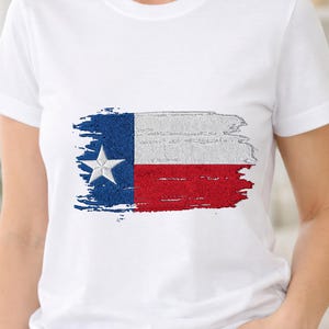 Puede incluir: Camiseta blanca con un diseño bordado de la bandera de Texas. El diseño de la bandera incluye una franja vertical azul con una estrella blanca, una franja horizontal blanca y una franja horizontal roja. El diseño de la bandera tiene un efecto de pincelada desgastada.