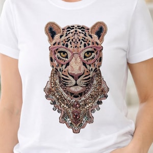 Puede incluir: Camiseta blanca con un diseño de leopardo bordado. El leopardo lleva gafas rosas y un collar de cuentas. El pelaje del leopardo es marrón con manchas negras. El diseño es detallado y llamativo.