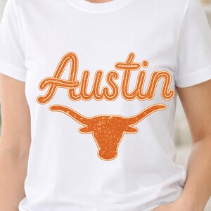 Peut inclure: T-shirt blanc avec le mot "Austin" en orange, écrit en script au-dessus d'une silhouette de longhorn orange. Le design a un aspect vieilli et vintage. Le t-shirt est à col rond et à manches courtes.