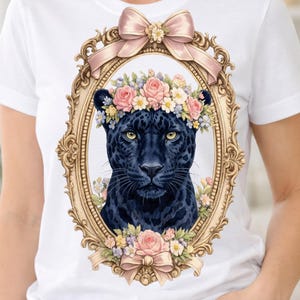Puede incluir: Camiseta blanca con una ilustración detallada de una pantera negra con una corona floral dentro de un marco dorado ornamentado. El diseño incluye lazos y flores rosas, creando una estética caprichosa y elegante. La camiseta es una prenda casual.