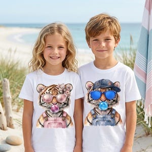 Puede incluir: Dos niños con camisetas blancas con gráficos de tigre. El tigre de la izquierda lleva gafas de sol rosas, una flor y un peto rosa. El tigre de la derecha lleva gafas de sol azules, una gorra y un peto vaquero. Ambos tigres están soplando burbujas.