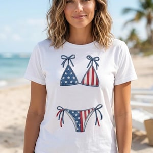 Puede incluir: Una camiseta blanca de manga corta con un estampado gráfico de un bikini de dos piezas. La parte superior e inferior del bikini están decoradas con un patrón de estrellas azules y blancas en un lado y rayas verticales rojas y blancas en el otro.
