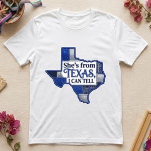 Puede incluir: Una camiseta blanca de manga corta con un gráfico del estado de Texas en un patrón de patchwork azul y plateado. El texto "She's from Texas, I CAN TELL" está impreso en letras azul oscuro en el centro de la forma del estado.