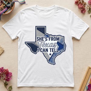 Puede incluir: Una camiseta blanca de manga corta con un gráfico del estado de Texas en azul marino y purpurina plateada. El diseño incluye el texto "SHE'S FROM Texas, I CAN TELL" en letras blancas y azul marino centradas dentro del contorno del estado.