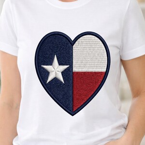 Puede incluir: Camiseta blanca con un bordado en forma de corazón con la bandera del estado de Texas. El diseño incluye una estrella blanca sobre fondo azul, una franja blanca y una franja roja. La camiseta está hecha de un material suave y cómodo.