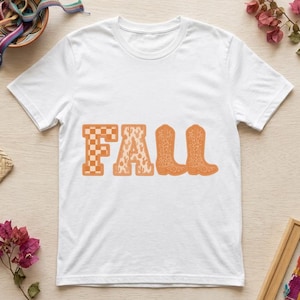 Puede incluir: Una camiseta blanca de manga corta con la palabra FALL impresa en naranja. La letra F tiene un patrón de cuadros, la letra A un estampado de cactus y las letras L y L tienen forma de dos botas vaqueras de color naranja.