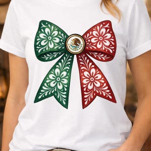 May include: White t-shirt featuring a large bow design in green and red, adorned with white floral patterns. The bow's center showcases a circular emblem with a detailed illustration. The shirt is a casual wear item.