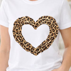 Puede incluir: Camiseta blanca con un diseño de corazón grande. El corazón está delineado con un patrón de estampado de leopardo marrón y negro. La camiseta tiene mangas cortas y cuello redondo.