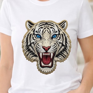 Puede incluir: Camiseta blanca con un diseño de cabeza de tigre bordada. El tigre tiene rayas negras, ojos azules y una boca roja con dientes visibles. El bordado está delineado en dorado.