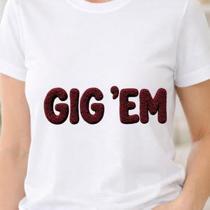 Könnte beinhalten: Weißes T-Shirt mit dem Text "GIG 'EM" in strukturiertem, weinrotem Farbton. Das T-Shirt hat kurze Ärmel und scheint aus einem weichen Material zu sein. Der Text ist mittig auf der Brust platziert.