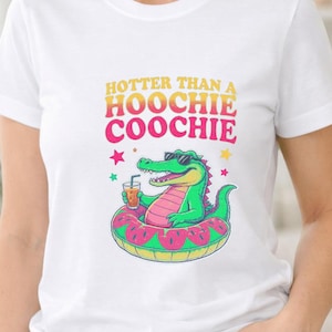 May include: A white short-sleeved t-shirt featuring a graphic of a green alligator wearing sunglasses, sitting in a pink and green watermelon-patterned pool float while holding a drink. The text reads HOTTER THAN A HOOCHIE COOCHIE in yellow and pink.