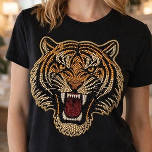Puede incluir: Camiseta negra con un diseño de cara de tigre. La cara del tigre está hecha de pequeñas cuentas doradas, naranjas, negras y blancas. La boca del tigre está abierta, revelando detalles rojos.