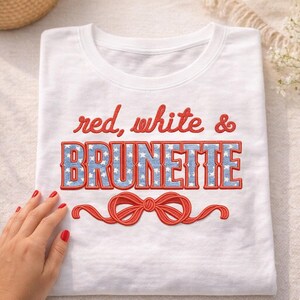 Puede incluir: Camiseta blanca con la frase bordada "red, white & BRUNETTE". La palabra "BRUNETTE" es azul con estrellas blancas, y el resto del texto es rojo. Un diseño de lazo rojo está debajo del texto.