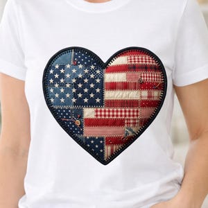 Puede incluir: Una camiseta blanca de manga corta con un gran parche en forma de corazón en el pecho. El parche presenta un diseño de patchwork que combina tela azul con estrellas blancas y patrones de tela a rayas o cuadros en rojo y blanco.