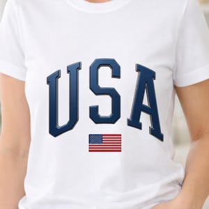 Puede incluir: Una camiseta blanca de manga corta que presenta las letras USA en una fuente grande, negrita y azul oscuro con un efecto biselado. Debajo del texto hay un pequeño gráfico rectangular de la bandera de los Estados Unidos con sus características rayas rojas, blancas y azules.