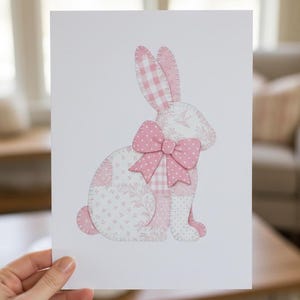 Puede incluir: Ilustración de un conejo rosa y blanco sobre fondo blanco. El conejo está hecho de tela de patchwork con varios patrones, incluyendo cuadros, lunares y diseños florales. Un gran lazo rosa adorna el cuello del conejo. La imagen es sostenida por una mano.