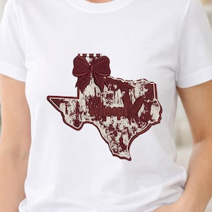 May include: White t-shirt featuring an embroidered design of the state of Texas in maroon and cream colors. The design includes a bow, the word "Whoop!", and a heart shape.