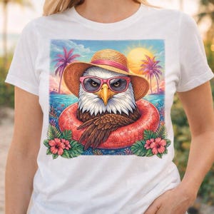 Puede incluir: Camiseta blanca con un águila de dibujos animados con gafas de sol y un sombrero de paja, flotando en un flotador rojo. El águila está situada en una escena de playa tropical con palmeras y una puesta de sol.