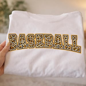 Puede incluir: Camiseta blanca doblada con la palabra "BASEBALL" en un diseño audaz y arqueado. Las letras presentan un estampado de leopardo con un contorno dorado. El diseño está centrado.