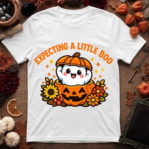 Puede incluir: Una camiseta blanca con el gráfico de un fantasma sonriente saliendo de una calabaza de Halloween tallada, rodeado de girasoles y hojas de otoño. El texto "EXPECTING A LITTLE BOO" está impreso en naranja sobre la ilustración central.
