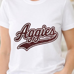 May include: White t-shirt with the word "Aggies" in a maroon glitter script. The word is outlined in a darker maroon, creating a striking design. The shirt is a classic crew neck style, suitable for casual wear.