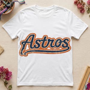 Puede incluir: Una camiseta blanca de manga corta con la palabra Astros impresa en el pecho en una fuente azul marino desgastada con un borde naranja grueso. El gráfico tiene una textura desgastada de estilo vintage en las letras.