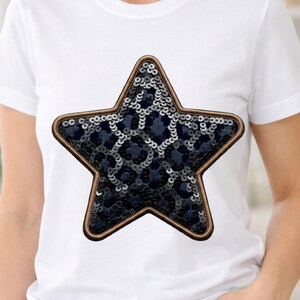 Puede incluir: Camiseta blanca con una gran aplicación de estrella de lentejuelas. La estrella tiene un centro azul oscuro estampado con lentejuelas plateadas y un borde marrón. La camiseta es de manga corta y probablemente de algodón.