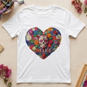 Puede incluir: Una camiseta blanca de manga corta con un gran gráfico en forma de corazón en el pecho. El corazón contiene una colorida calavera de azúcar rodeada de intrincados patrones florales en tonos rojo, azul, amarillo y verde, con la palabra MEXICO impresa debajo.