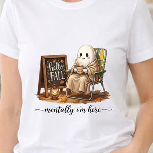Puede incluir: Una camiseta blanca con un gráfico de un fantasma envuelto en una manta beige, sentado en una silla plegable con una taza de café. Junto al fantasma hay una pizarra que dice hello FALL, una linterna y hojas de otoño caídas.