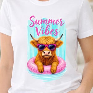 Puede incluir: Una camiseta blanca con un gráfico de una vaca Highland marrón con gafas de sol rosas. La vaca está dentro de un flotador inflable rosa, sobre un fondo de rayas verticales azul claro con el texto Summer Vibes en letra cursiva rosa.