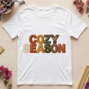 Puede incluir: Una camiseta blanca de manga corta con el texto COZY SEASON impreso en el centro. Las letras están diseñadas para parecer parches de tela en varios colores y texturas inspirados en el otoño, incluyendo marrón, naranja, verde y patrones de cuadros.