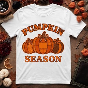 Puede incluir: Una camiseta blanca de manga corta con un gráfico de tres calabazas naranjas con patrones de cuadros y lazos. El texto PUMPKIN SEASON está impreso en letras de bloque naranjas y marrones sobre y debajo de la ilustración en el frente.