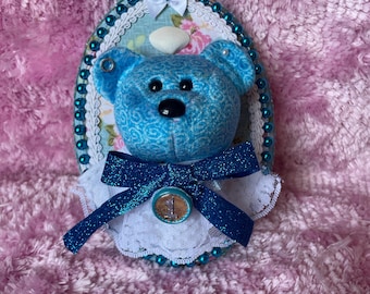 May include: An oval-shaped wall plaque featuring a blue patterned teddy bear head with black eyes and nose. The bear wears a white lace collar and a dark blue glittery bow with a circular button displaying the number 1. The plaque is bordered by teal beads.